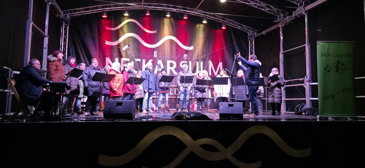 Sing'n'Fun singt abends auf der Bühne des Neckarsulmer Weihnachtsmarkts, daneben das Banner des Chors